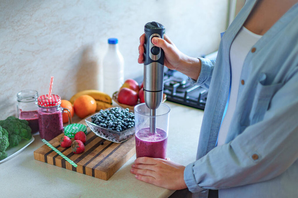 Woman cooking fresh blueberry smoothie using hand blender at kitchen at home. Healthy eating  - Foto, imagen
