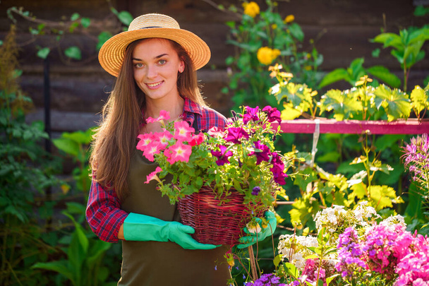 Portrait of happy joyful florist gardener woman in hat with flower pot of petunia outdoors. Gardening and floriculture. Growing flower in home garden  - Fotó, kép