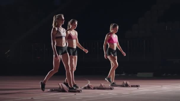 A row of runners womens crouch in the starting position before beginning to race. Females start with running shoes on the stadium from the start line in the dark with spotlights in slow motion. - Záběry, video