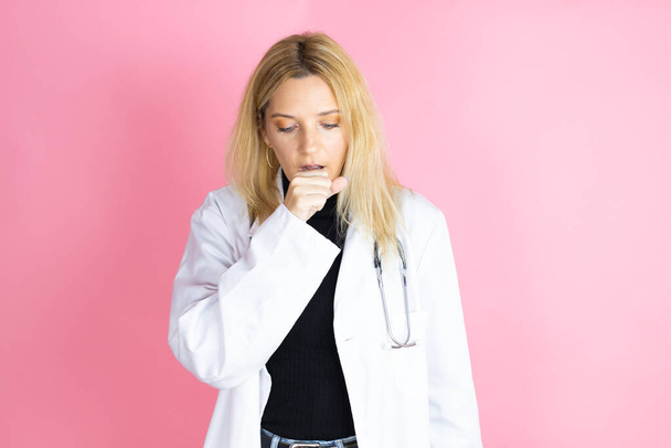 Young blonde doctor woman wearing stethoscope standing over isolated pink background with her hand to her mouth because she's coughing - Photo, Image