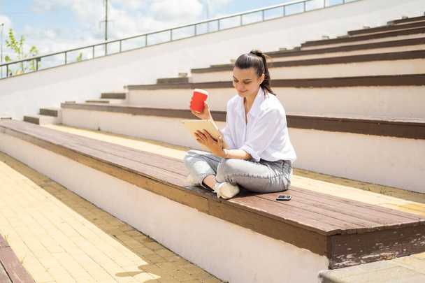 Счастливые молодые женщины с планшетом держа чашку кофе бумагу наслаждаясь солнечным днем сидя в амфитеатре. - Фото, изображение