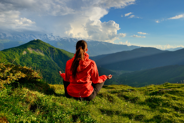 Jeune femme en veste rouge assis dans la pose de yoga dans les montagnes
 - Photo, image