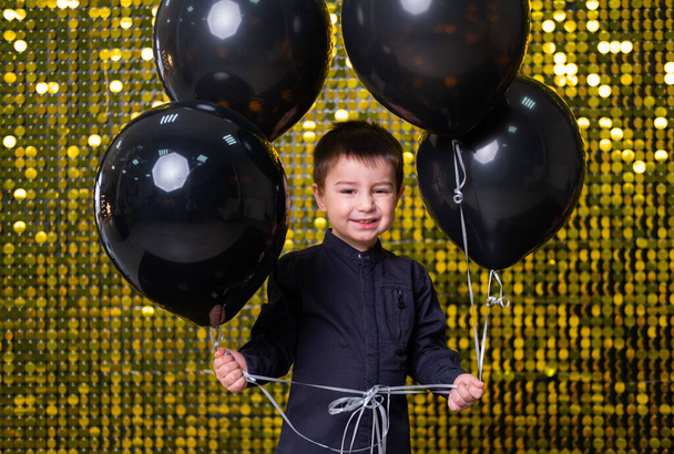chłopiec trzyma czarne balony na tle ze złotymi, błyszczącymi cekinami, pailletkami. - Zdjęcie, obraz