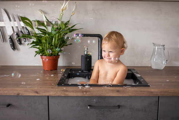 Cute happy baby girl with playing with water and foam in a kitchen sink at home - Photo, Image