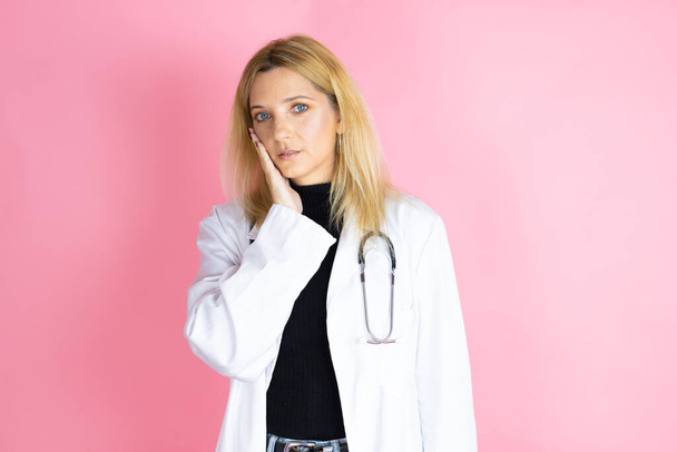 Young blonde doctor woman wearing stethoscope standing over isolated pink background touching mouth with hand with painful expression because of toothache or dental illness on teeth - 写真・画像