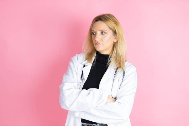 jonge blonde arts vrouw dragen stethoscoop staan over geïsoleerde roze achtergrond denken kijken moe en verveeld met gekruiste armen - Foto, afbeelding