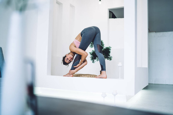 Full body of barefoot young woman in sport clothes doing stretching exercise while practicing yoga in light spacious living room - Valokuva, kuva