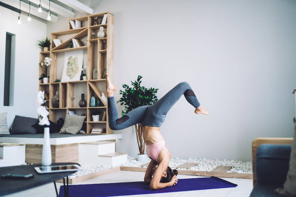 Full body of young slim focused anonymous woman in activewear practicing sirsasana pose while training alone on mat in light cozy room - Foto, Imagen