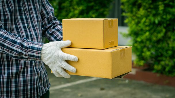 Młody dostawca w białych rękawiczkach trzymający pudełko przed domem, koncepcja dostawy online. - Zdjęcie, obraz
