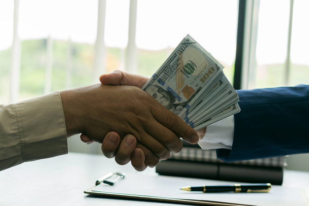 Два бизнесмена пожимают руки с большими долларами в руках после того, как либерализация разберется с коррупцией сверху. - Фото, изображение