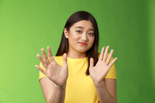 Modest cute insecure timid asian girl waving hands no gesture, step back, smirk, cringe aversion and dislike, refusing suspiciosu offer, rejecting proposal, stand green background - Фото, изображение