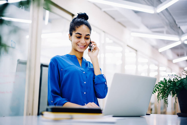 Positive young lady smiling while answering phone and using laptop in modern office while standing near table and wearing shirt - Zdjęcie, obraz