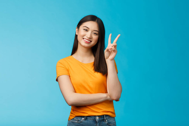 Optimistic cute tender asian young millennial girl send positive vibes, tilt head show peace, victory sign, smiling broadly, wish fantastic holidays, stand blue background joyful upbeat - Photo, Image