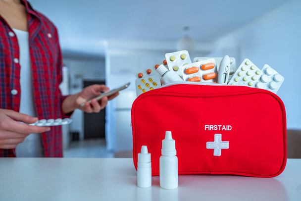 Woman using smartphone for reading and examining medicine instruction on the internet before take pills. First aid kit on table at home - Photo, Image