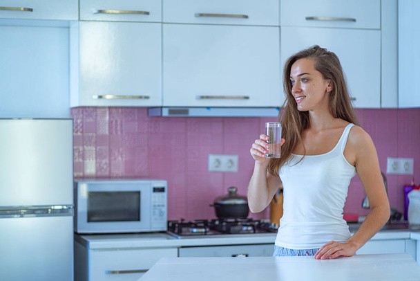 Ευτυχισμένη νυσταγμένη χαρούμενη γυναίκα πίνει ένα ποτήρι καθαρό καθαρό πρωινό νερό νωρίς το πρωί αφού ξυπνήσει στην κουζίνα στο σπίτι. Υγιεινός τρόπος ζωής Αρχή και έναρξη μιας νέας καλής ημέρας  - Φωτογραφία, εικόνα
