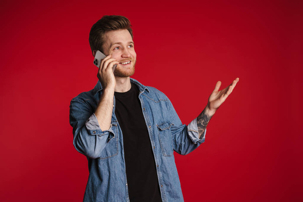 Happy smiling young man in casual clothes standing over red wall background, holding mobile phone, calling - Fotoğraf, Görsel