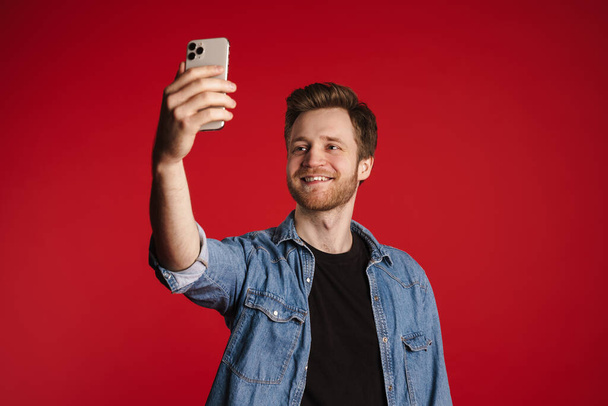 Happy smiling young man in casual clothes standing over red wall background, taking a selfie - Fotó, kép