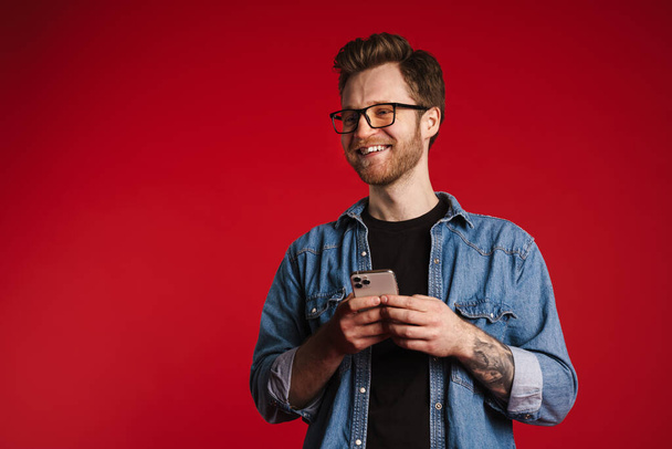 Happy smiling young man in casual clothes standing over red wall background, holding mobile phone - Foto, imagen