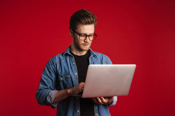 Calm young man in casual clothes standing over red wall background, holding laptop computer - Фото, зображення