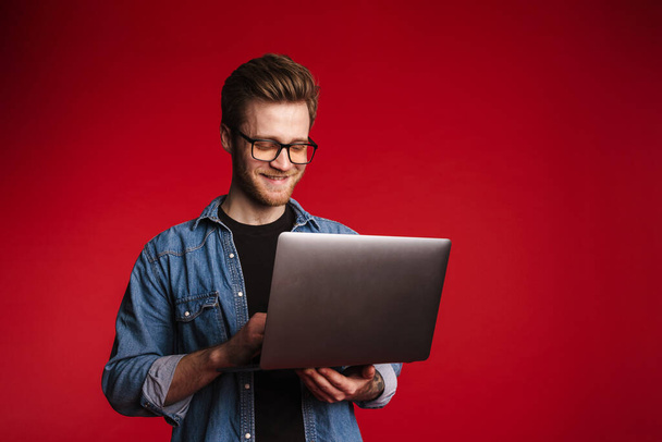Happy smiling young man in casual clothes standing over red wall background, holding laptop computer - Foto, Bild