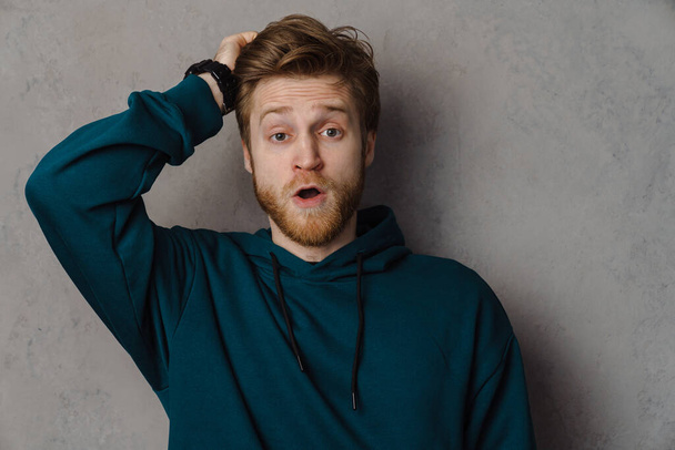 Bearded ginger man holding his head while expressing surprise isolated over grey background - Photo, Image