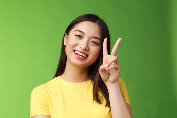 Közelkép vidám aranyos aranyos ázsiai lány élénk megjelenés kamera mosoly, mutatják béke győzelem jele szórakoztatott, vigyorgó pózol fénykép gyengéd kifejezés, állni zöld háttér pozitív és szerencsés - Fotó, kép