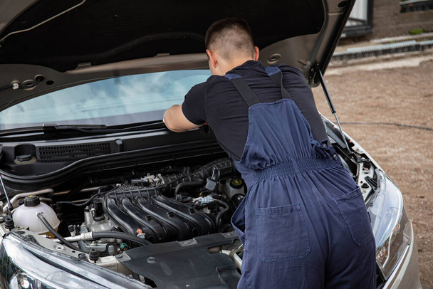 a man repairs a car. a man in blue special clothes to repair the car. engine parts machine repair maintenance breakage problem hands people man master - Фото, зображення