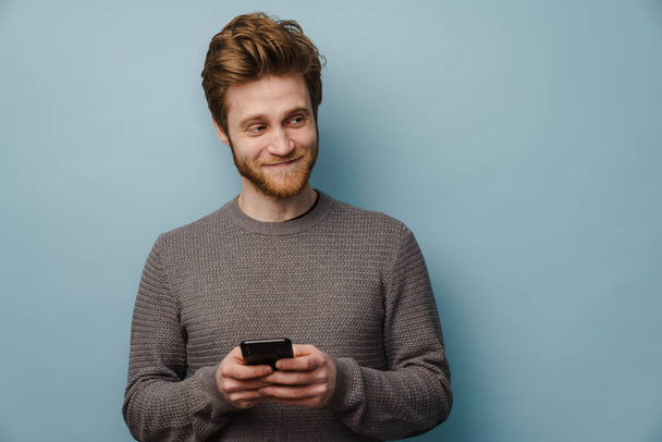 White ginger man with beard smiling and using mobile phone isolated over blue background - Φωτογραφία, εικόνα