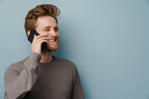 White ginger man with beard smiling while talking on mobile phone isolated over blue background - Valokuva, kuva