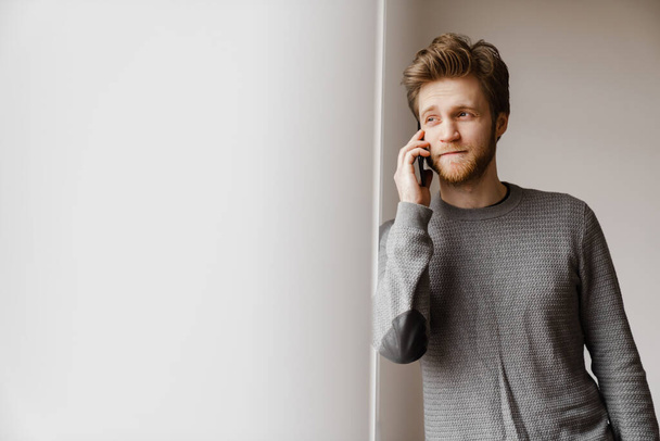 Ginger young man talking on mobile phone while standing indoors - Фото, изображение