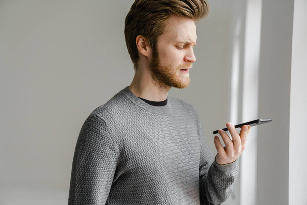 Ginger young man talking on mobile phone while standing indoors - Fotografie, Obrázek