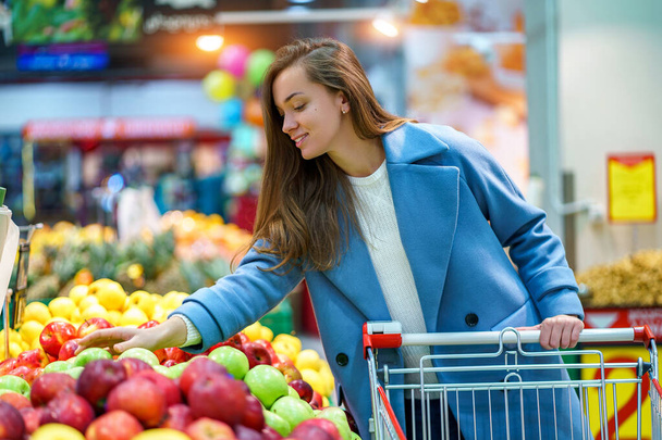 Portrait of a smiling happy attractive young woman buyer with cart in the grocery supermarket during choosing and buying fresh apples at fruit department - Foto, imagen