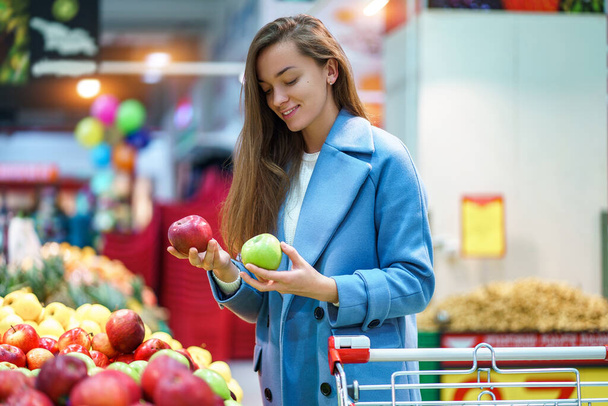 Portrait of attractive woman buyer with cart in the grocery shop during choosing and buying fresh apples at fruit department - Foto, imagen