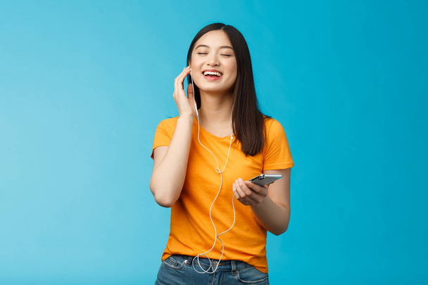 Lovely ξέγνοιαστη χαριτωμένο κορίτσι της Ασίας απολαύσετε φοβερό μουσική ώθηση διάθεση ακούγοντας αγαπημένα τραγούδια, αγγίξτε ακουστικό κοντά μάτια από απόλαυση ευχαρίστηση, τραγουδώντας μαζί, κρατήστε το smartphone σταθεί μπλε φόντο - Φωτογραφία, εικόνα