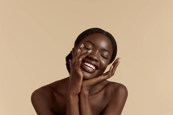 Portrait close up of beautiful african girl with closed eyes. Smiling young woman touch her clean face. Concept of face skin care. Isolated on beige background. Studio shoot - Fotoğraf, Görsel