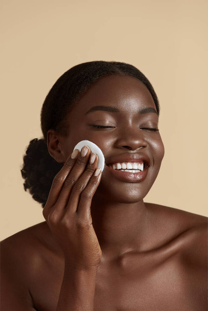 Portrait close up of beautiful black girl touch her clean face with cotton pad. Smiling young woman with closed eyes. Concept of face skin care. Isolated on beige background. Studio shoot - Foto, imagen