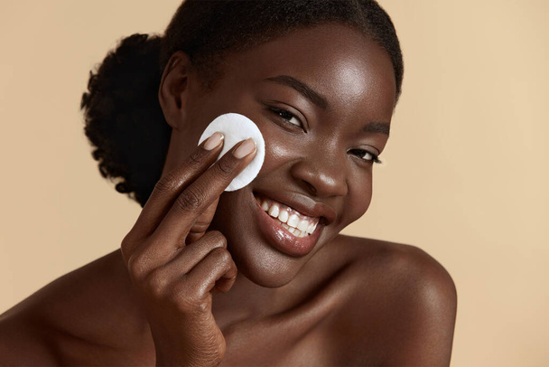 Portrait close up of beautiful black girl touch her clean face with cotton pad. Smiling young woman looking at camera. Concept of face skin care. Isolated on beige background. Studio shoot - Foto, afbeelding