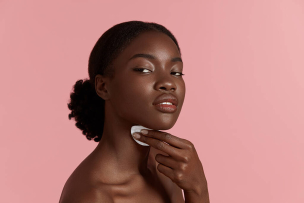 Portrait close up of beautiful black girl touch her clean face with cotton pad. Serious young woman. Concept of face skin care. Isolated on pink background. Studio shoot - Photo, image