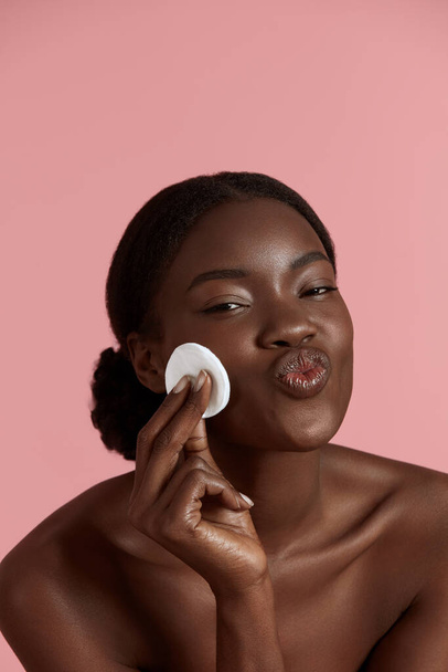 Portrait close up of beautiful black girl touch her clean face with cotton pad. Smiling young woman show her lips. Concept of face skin care. Isolated on pink background. Studio shoot - Foto, afbeelding