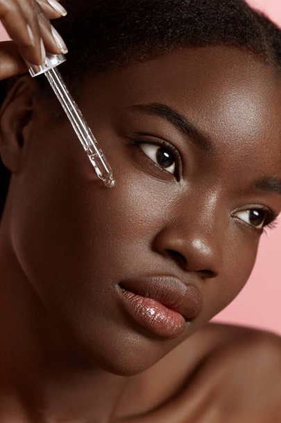 Portrait close up of beautiful black girl dropping serum collagen moisturizer on face. Serious young woman. Concept of face skin care. Isolated on pink background. Studio shoot - Fotoğraf, Görsel
