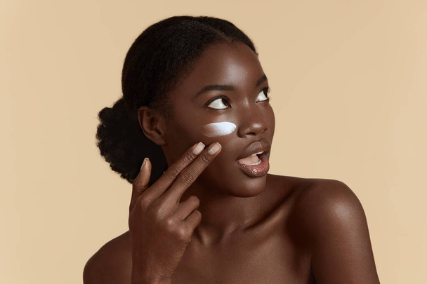 Portrait close up of beautiful black girl apply cosmetic cream on face. Surprised young woman with perfect skin. Concept of face skin care. Isolated on beige background. Studio shoot - 写真・画像