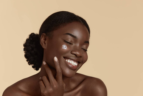 Portrait close up of beautiful black girl with cosmetic cream on face. Smiling young woman with closed eyes. Concept of face skin care. Isolated on beige background. Studio shoot - Photo, Image