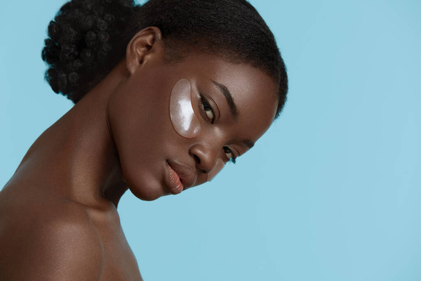 Close up portrait of beautiful african girl with under eye patch on face. Serious young woman looking at camera. Concept of face skin care. Isolated on blue background. Studio shoot - Zdjęcie, obraz
