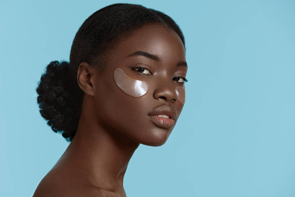 Close up portrait of pretty african girl with under eye patch on face. Serious young woman with perfect skin looking at camera. Concept of face skin care. Isolated on blue background. Studio shoot - Photo, image