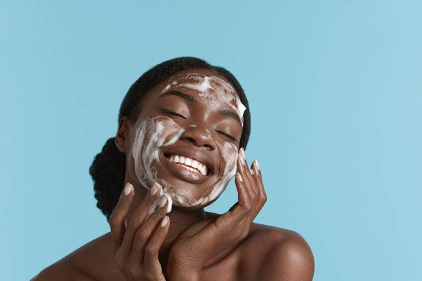 Close up portrait of beautiful black girl wash her face with cleansing face foam. Smiling young woman with closed eyes. Concept of face skin care. Isolated on blue background. Studio shoot - Foto, Bild