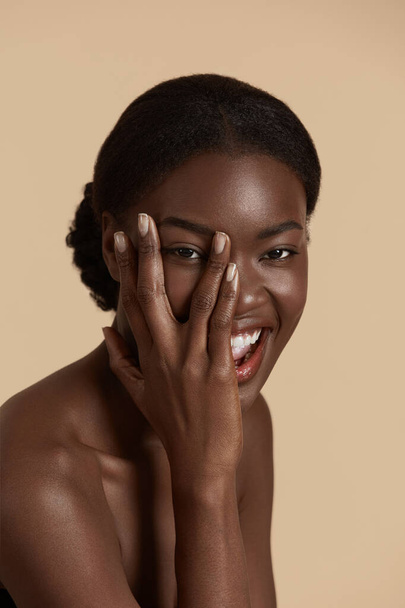 Portrait close up of beautiful african girl. Smiling young woman touch her clean face and looking at camera. Concept of face skin care. Isolated on beige background. Studio shoot - Photo, image