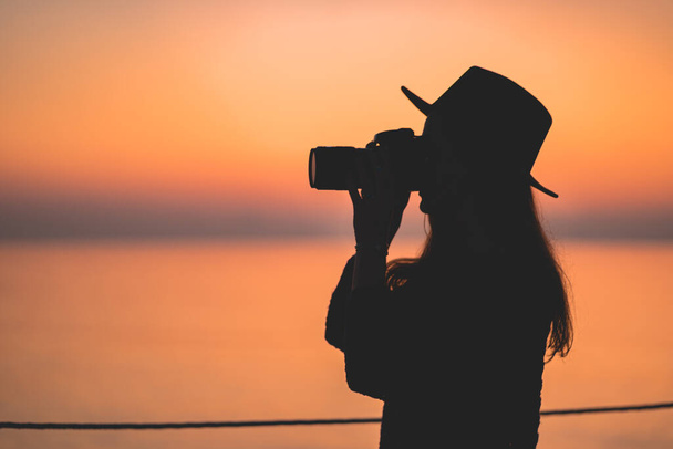 Silhouette of a woman photographer in hat with dslr camera during taking photos at sunset - Zdjęcie, obraz