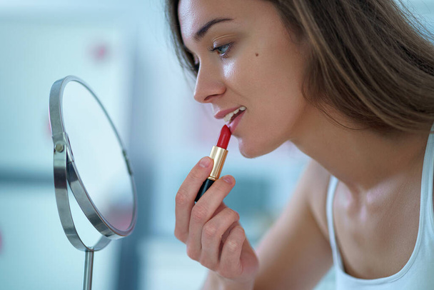 Beautiful attractive happy smiling brunette woman paints lips with red lipstick using a small round mirror during home makeup in the morning - Foto, afbeelding