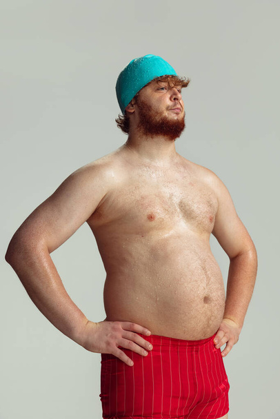 Funny cheerful swimmer. Cute red-headed man in red swimming shorts posing isolated on gray studio background. Concept of sport, humor and body positive. - Foto, Bild
