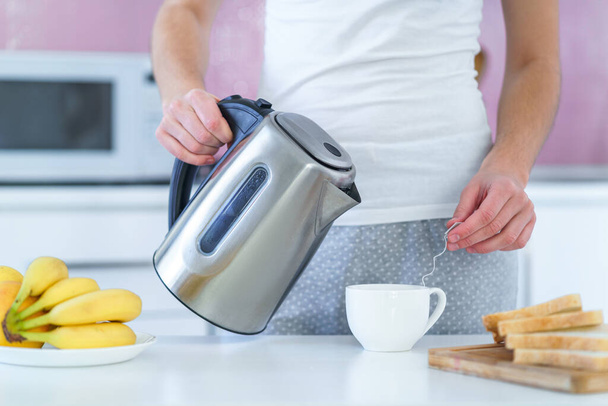 Mujer vertiendo agua caliente de hervidor eléctrico para preparar té para la hora del té en la cocina en casa - Foto, imagen
